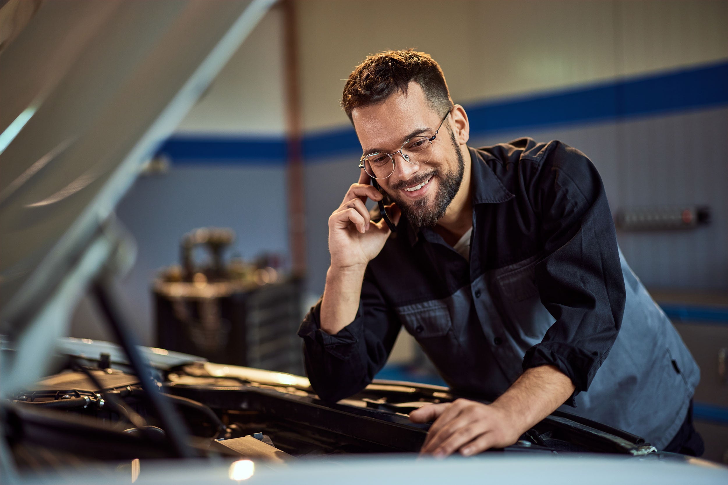 Mechanic providing support on a phone next to an open car hood in a garage.